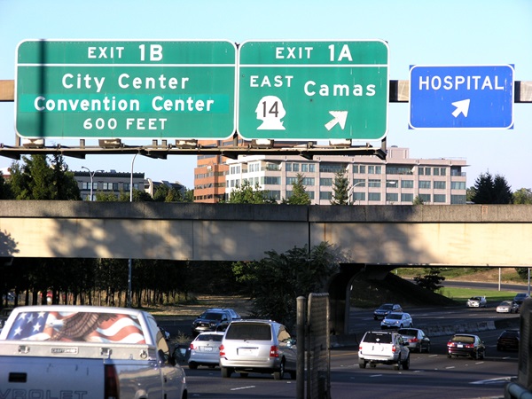 Cars driving on a busy freeway beneath large green overhead signs for Exit 1B to City Center and Convention Center, Exit 1A to Camas on State Route 14 East, and a blue hospital exit sign, with office buildings and trees visible in the background.